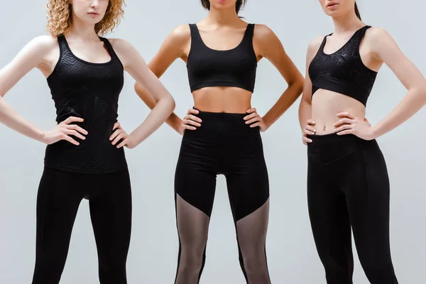 Vue recadrée de femmes multiculturelles en vêtements de sport debout avec les mains sur les hanches isolées sur blanc — Photo de stock