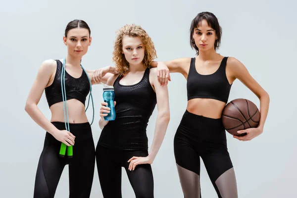 Chicas atractivas y multiculturales de pie con equipos deportivos aislados en blanco - foto de stock