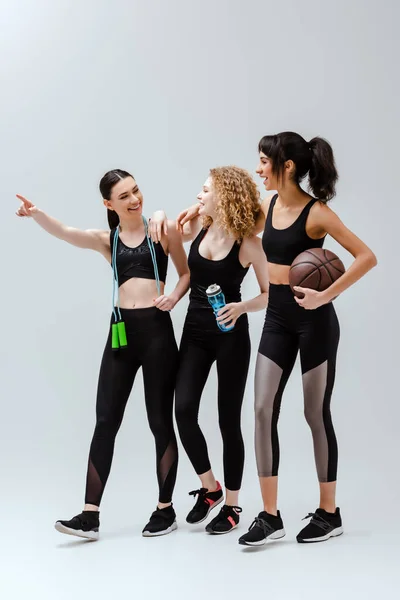 Cheerful woman pointing with finger near multicultural girls walking with sport equipment on white — Stock Photo