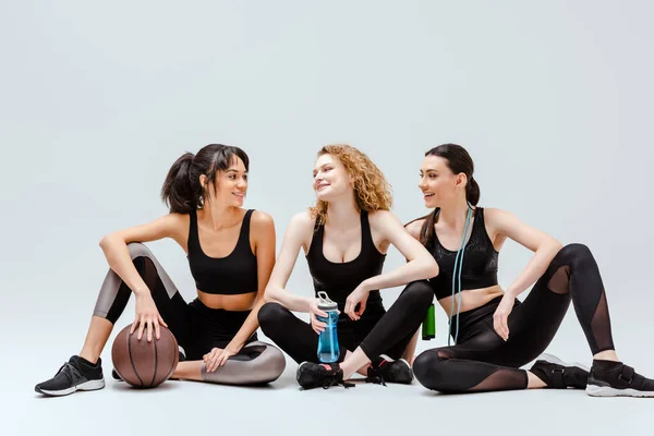 Fröhliche multikulturelle Mädchen sitzen mit Sportgeräten auf weiß — Stockfoto