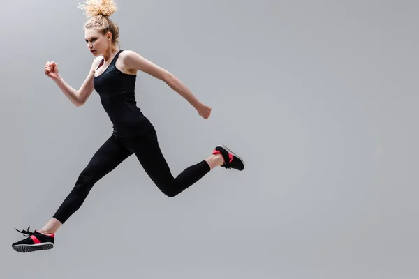 Молода і кучерява спортсменка в спортивному одязі стрибає ізольовано на сірому — стокове фото