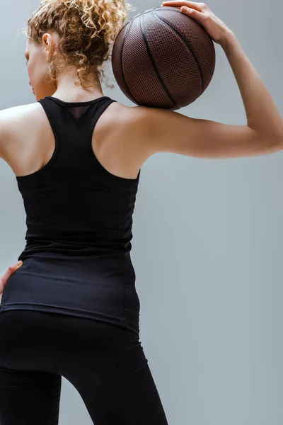 Кучерява молода спортсменка в спортивному одязі тримає баскетбол ізольовано на сірому — стокове фото
