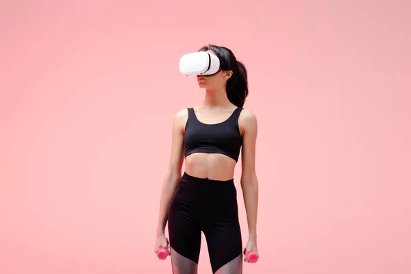 African american sportswoman in virtual reality headset holding dumbbells on pink — Stock Photo