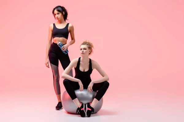 Ragazza sportiva seduta sul centro commerciale fitness vicino alla donna americana africana con bottiglia sportiva sul rosa — Foto stock