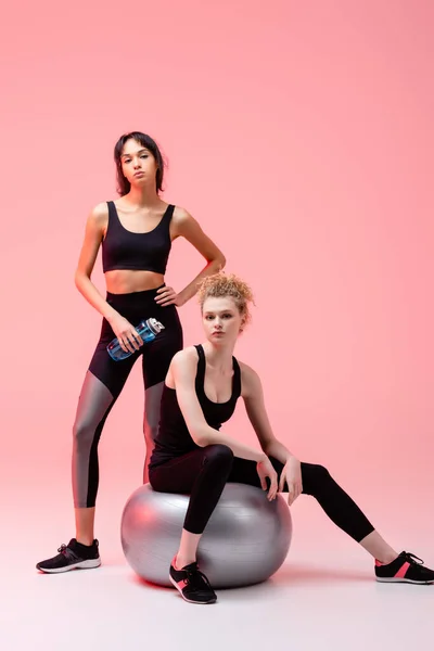 Chica deportiva sentada en el centro comercial de fitness cerca de la deportista afroamericana con botella de deportes posando en rosa - foto de stock