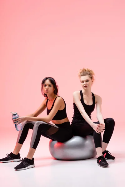 Fille bouclée assis sur le centre commercial de remise en forme avec une femme afro-américaine tenant bouteille de sport sur rose — Photo de stock