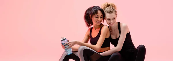 Plan panoramique de fille heureuse assise sur le centre commercial de remise en forme avec femme afro-américaine gaie isolé sur rose — Photo de stock