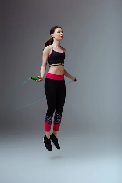 Привлекательная женщина в спортивной одежде прыгает с скакалкой на сером — стоковое фото