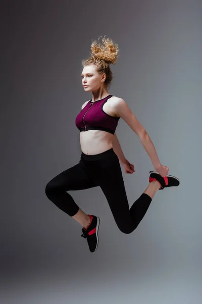 Кучерява і спортивна жінка в спортивному одязі стрибає на сірому — стокове фото