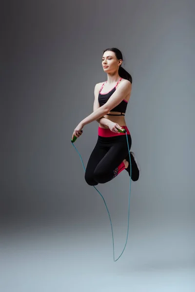 Красива жінка в спортивному одязі стрибає з пропусканням мотузки на сірому — стокове фото