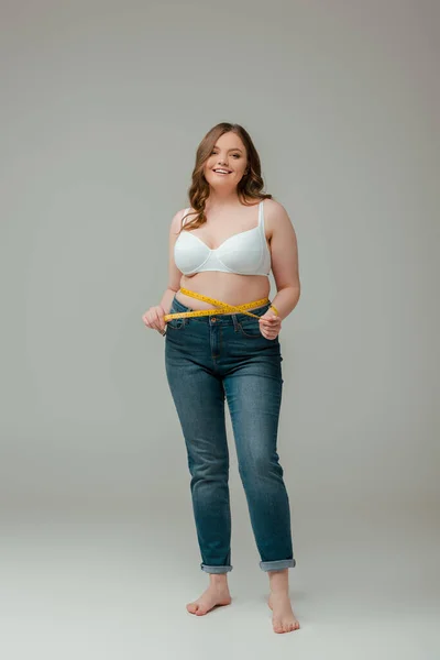 Happy plus size woman in jeans and bra measuring waist on grey — Stock Photo