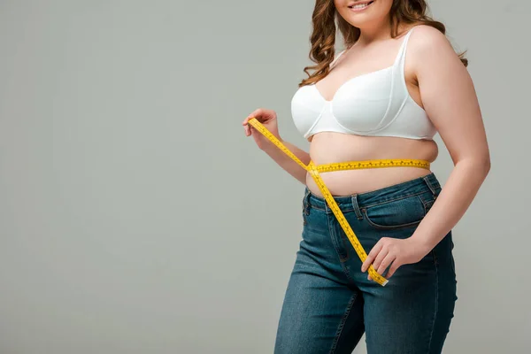 Cropped view of happy plus size woman in jeans measuring waist isolated on grey — Stock Photo