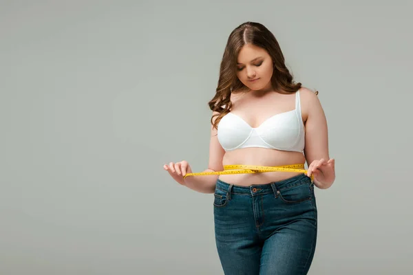 Plus size girl in jeans measuring waist isolated on grey — Stock Photo