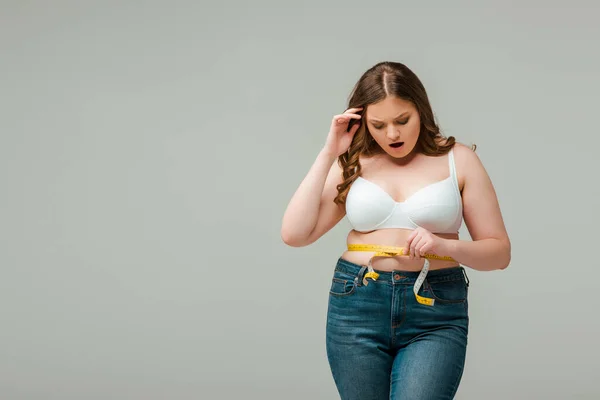 Shocked plus size woman in jeans measuring waist isolated on grey — Stock Photo