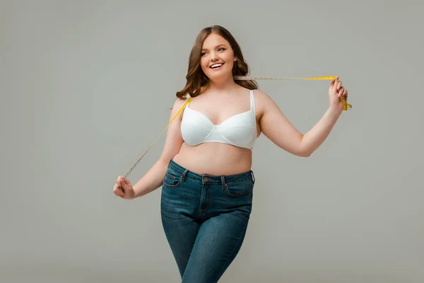 Счастливая плюс размер женщина в джинсах проведение измерительной ленты изолированы на серый — стоковое фото