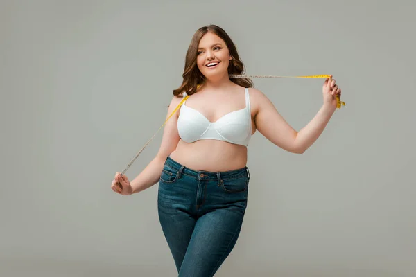 Happy plus size woman in jeans holding measuring tape isolated on grey — Stock Photo