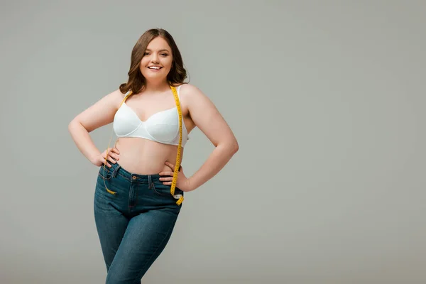 Happy plus size woman in jeans and bra standing with hands on hips isolated on grey — Stock Photo
