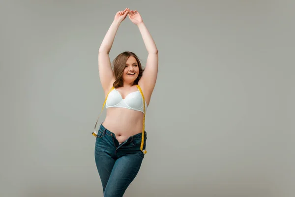 Felice plus size donna in jeans e reggiseno in piedi con le mani sopra la testa isolato su grigio — Foto stock