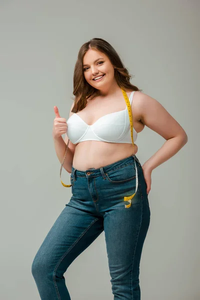 Cheerful plus size woman in jeans and bra standing with measuring tape and showing thumb up isolated on grey — Stock Photo