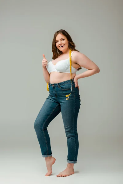 Cheerful plus size woman in jeans and bra standing with measuring tape and showing thumb up on grey — Stock Photo