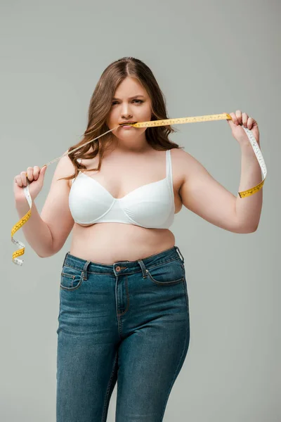 Além de tamanho mulher em jeans e sutiã mordendo fita métrica isolada em cinza — Fotografia de Stock