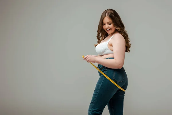 Cheerful plus size woman in jeans and bra measuring body isolated on grey — Stock Photo