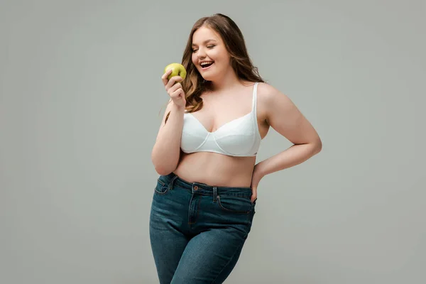 Cheerful plus size woman in jeans and bra holding apple isolated on grey — Stock Photo