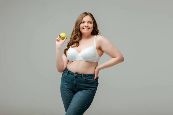Happy plus size woman in jeans and bra holding apple isolated on grey — Stock Photo
