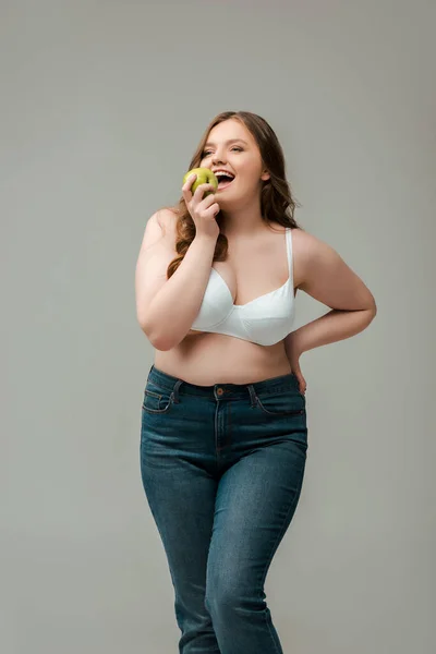 Cheerful plus size woman in jeans and bra eating apple isolated on grey — Stock Photo