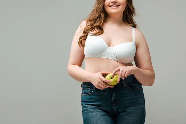 Vista ritagliata di allegra donna plus size in jeans e reggiseno con mela isolato su grigio — Foto stock