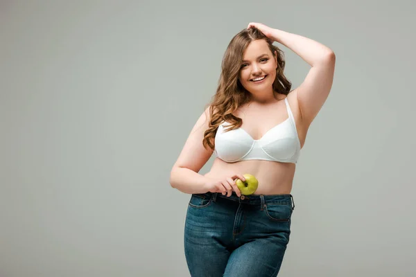 Счастливая плюс размер девушка в джинсах и бюстгальтере держа яблоко изолированы на серый — стоковое фото