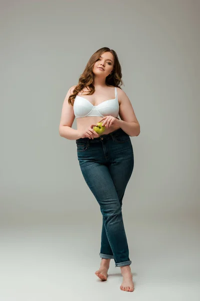 Приваблива дівчина плюс розмір в джинсах і бюстгальтер тримає яблуко на сірому — стокове фото