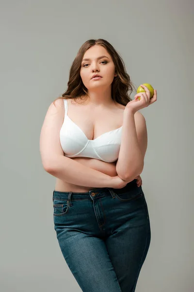 Attractive plus size girl in jeans and bra holding apple isolated on grey — Stock Photo