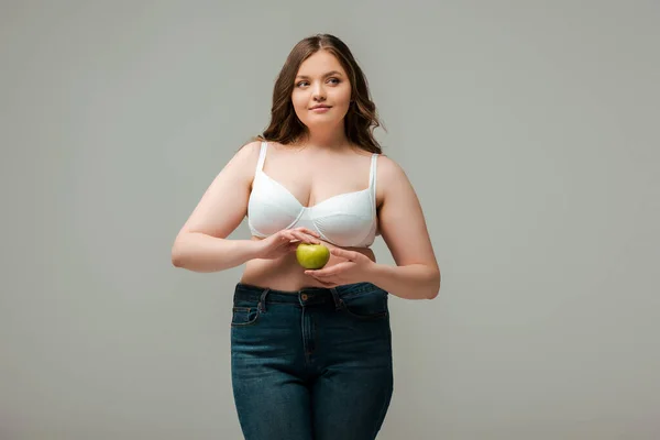 Plus size girl in jeans and bra holding apple isolated on grey — Stock Photo