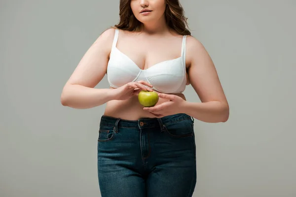 Cropped view of plus size girl in jeans and bra holding apple isolated on grey — Stock Photo