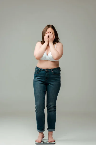Plus size woman in jeans and bra standing on scales and covering face on grey — Stock Photo