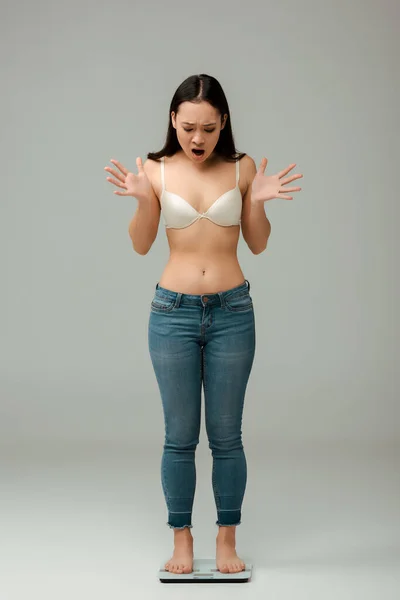 Scioccato asiatica in sovrappeso ragazza in jeans e reggiseno in piedi su scale e gesticolando sul grigio — Foto stock