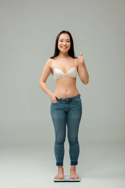 Happy overweight asian woman standing on scales and showing thumb up on grey — Stock Photo