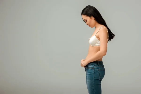 Side view of displeased and overweight asian girl wearing jeans isolated on grey — Stock Photo