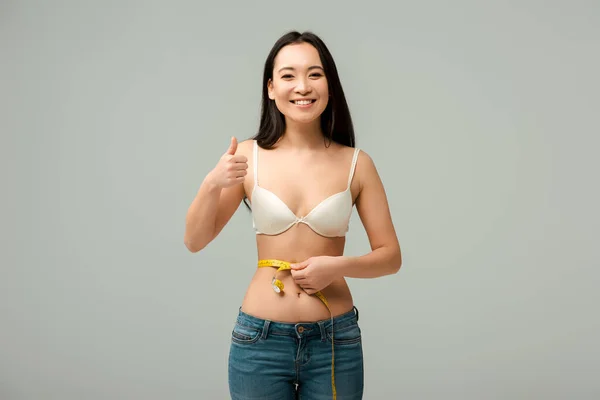 Счастливая и толстая азиатская девушка в бюстгальтере, измеряющая талию и показывающая большой палец, изолированный на сером — стоковое фото