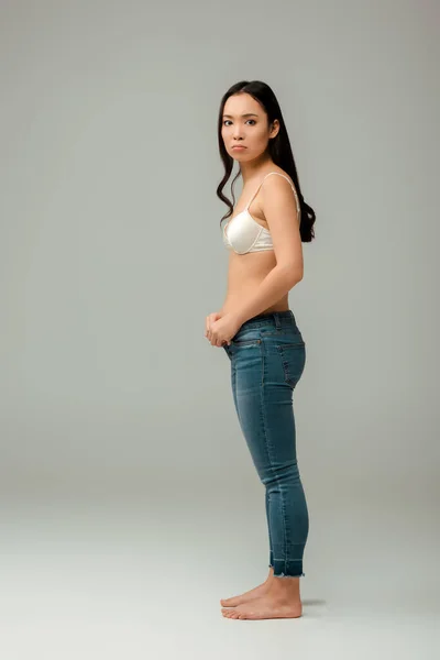 Disgustado y con sobrepeso mujer asiática usando jeans en gris - foto de stock