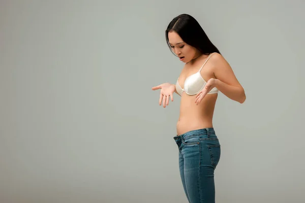 Mujer asiática disgustada y con sobrepeso haciendo gestos y mirando jeans aislados en gris - foto de stock