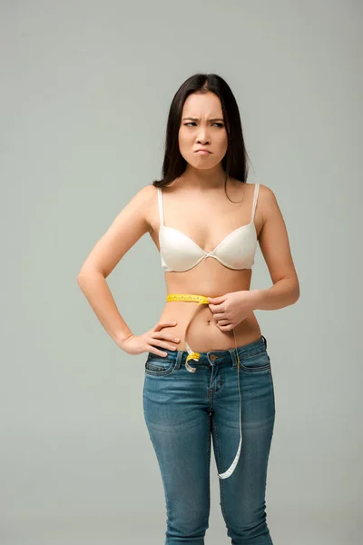 Upset and overweight asian girl measuring waist isolated on grey — Stock Photo