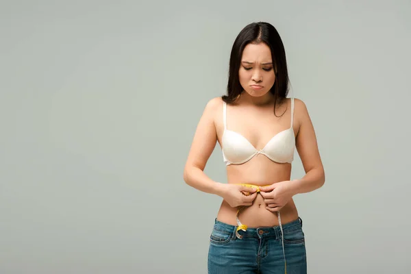 Незадоволена і надмірна вага азіатська дівчина вимірює талію ізольована на сірому — стокове фото