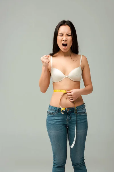 Emotional and overweight asian girl measuring waist and screaming isolated on grey — Stock Photo