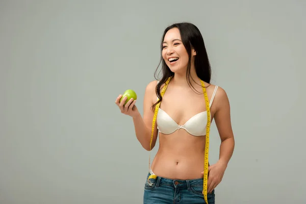 Щаслива і надмірна вага азіатська дівчина тримає мірну стрічку і яблуко ізольовано на сірому — стокове фото