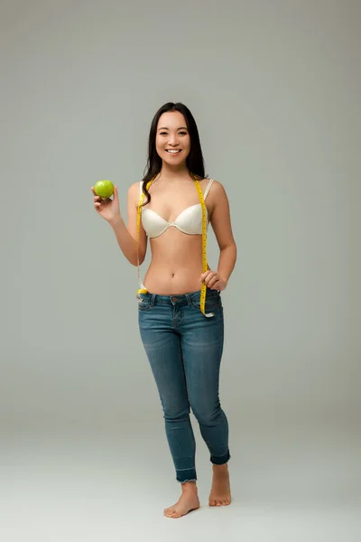 Happy and overweight asian girl holding measuring tape and apple on grey — Stock Photo