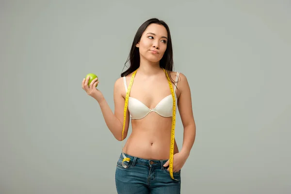 Недовольная азиатская девушка держит яблоко, стоя с рукой в кармане изолированы на серый — стоковое фото