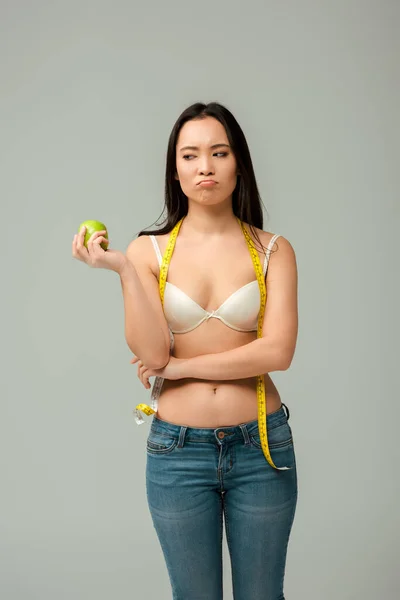 Незадоволена азіатська дівчина в бюстгальтері дивиться на яблуко ізольовано на сірому — стокове фото
