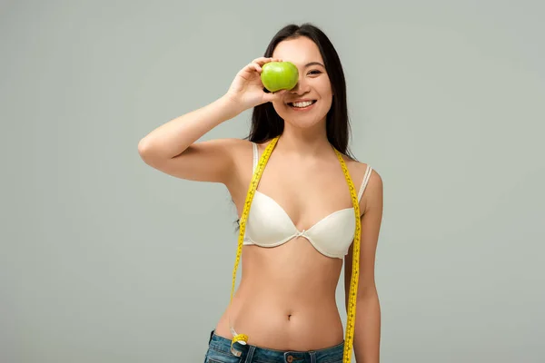 Улыбающаяся и толстая азиатская девушка, держащая яблоко рядом с лицом, изолированная на сером — стоковое фото